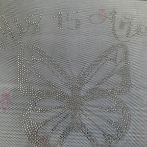 May include: A light gray fabric with a large butterfly design and the words "Mis 15 Años" spelled out in silver rhinestones. The number "15" is also in rhinestones. A small pink heart is visible.