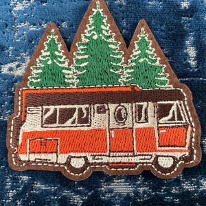 May include: An embroidered patch featuring a vintage orange and white camper van positioned beneath three green pine trees. The design is outlined in a dark brown border, showcasing detailed stitching of the vehicle windows, doors, and wheels.