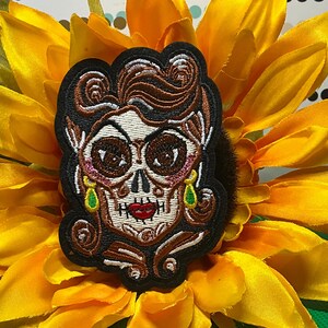 May include: Embroidered patch featuring a stylized sugar skull with brown hair, a black outline, and green teardrop earrings. The skull has large, dark eyes and a red mouth. The patch is set against a bright yellow sunflower.