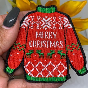 May include: Embroidered red Christmas sweater patch with the words "MERRY CHRISTMAS" in white. The patch features green reindeer, white snowflakes, and green and white trim. The patch is outlined in black.