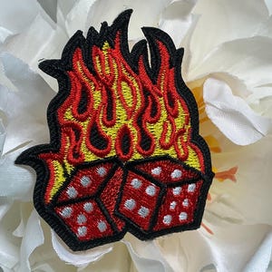 May include: An embroidered patch featuring two red dice with white dots positioned at the base of a stylized flame design. The flames are rendered in vibrant yellow and red thread, all outlined with a thick black border for high contrast.