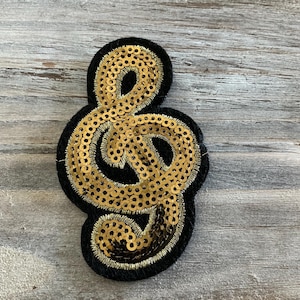 May include: A black and gold sequined treble clef patch. The musical note is outlined in black with gold sequins filling the shape. The patch is on a gray wooden surface.