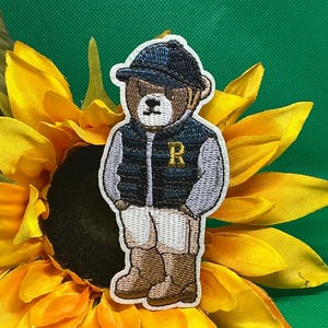 May include: Embroidered patch of a teddy bear wearing a navy blue vest with a gold "R", a matching cap, and khaki shorts. The bear is brown with a white outline. The patch is set against a yellow sunflower.