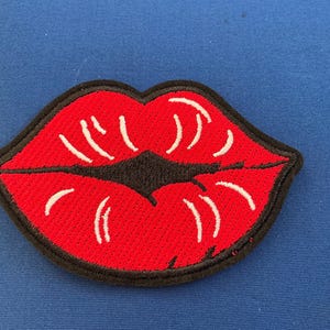May include: Embroidered patch featuring a pair of red lips with white highlights and a black outline. The lips are a vibrant red color, with a black area representing the mouth's opening. The patch is on a blue background.
