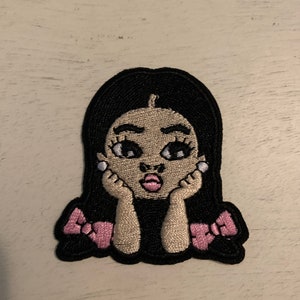 May include: An embroidered patch featuring a cartoon character with long black hair, tan skin, and pink lips. The character rests their chin on their hands and wears two pink bows in their hair. The design is outlined in black thread with white accents.