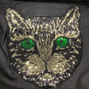 May include: A sequined cat face patch with bright green eyes. The cat's face is black with gold and silver sequins. The cat's eyes are a vibrant green color. The patch is likely for clothing or accessories.
