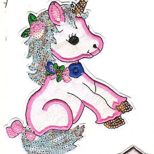 May include: A white unicorn with pink and blue accents, decorated with sequins and flowers. The unicorn has a pink mane and tail, a blue bow, and a gold horn.
