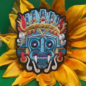 May include: Embroidered patch featuring a stylized blue face with intricate details, including a headdress and ornate facial features. The patch is set against a yellow sunflower with green background.