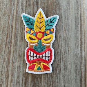 May include: Embroidered patch featuring a colorful Tiki mask design. The patch has a white border and showcases a yellow, red, brown, and teal color palette. The Tiki mask has a detailed design with a wide mouth and a crown of leaves.