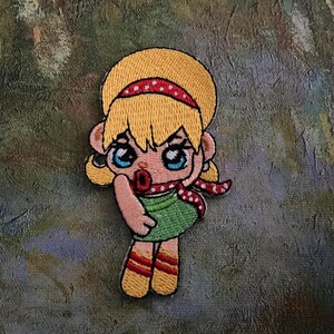 May include: Embroidered patch featuring a cartoon girl with blonde hair, blue eyes, and a red polka-dot headband and scarf. She wears a green dress and yellow socks and shoes. The patch is on a textured, multi-colored background.