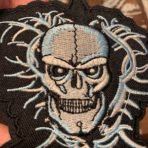 May include: A black embroidered patch with a white and light blue skull design. The skull has a detailed design with a cracked skull and a menacing expression. The skull is surrounded by a white and light blue design that resembles vines or thorns.