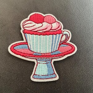 May include: An embroidered patch featuring a cupcake in a light blue and white striped cup, topped with pink frosting and two red cherries, resting on a red and blue pedestal cake stand with a white border and a white backing.