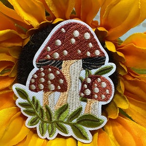 May include: An embroidered patch featuring three mushrooms with reddish-brown caps and white spots, set against a white background. The mushrooms have light-colored stems and are accented with green fern-like leaves at the base of the design.