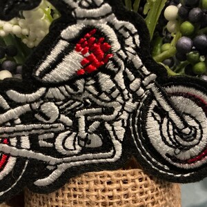Puede incluir: Parche bordado con un esqueleto montado en una motocicleta. El parche tiene un borde negro, con detalles blancos y rojos en las ruedas y el motor de la motocicleta. El pecho del esqueleto es rojo.