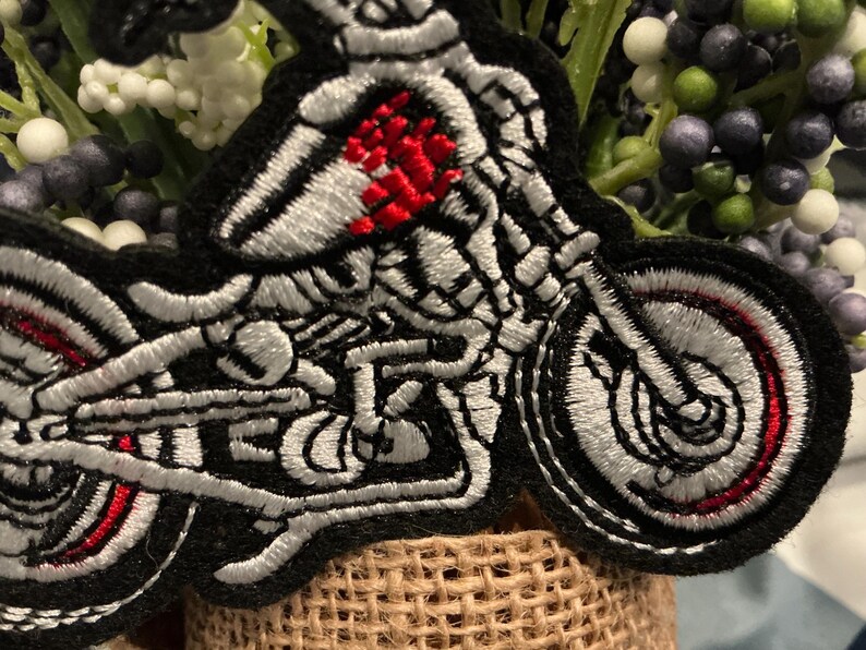 Puede incluir: Parche bordado con un esqueleto montado en una motocicleta. El parche tiene un borde negro, con detalles blancos y rojos en las ruedas y el motor de la motocicleta. El pecho del esqueleto es rojo.