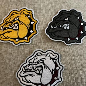 May include: Three embroidered patches featuring a stylized bulldog head in profile. One patch is yellow, one is black, and one is white. Each bulldog has a spiked collar, bared teeth, and a white border, showcasing detailed thread work.