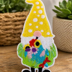 May include: An embroidered patch shaped like a gnome wearing a tall yellow hat with white polka dots. The gnome has a white beard, a pink nose, and black boots. It holds a colorful bouquet of flowers in shades of blue, red, and yellow.