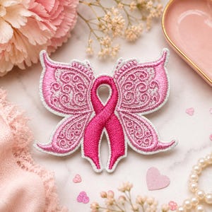May include: An embroidered patch featuring a bright pink awareness ribbon at the center, flanked by two symmetrical butterfly wings. The wings are detailed with intricate, swirling lace-like patterns in shades of pink and white thread.