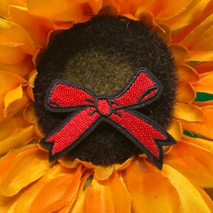 May include: A red bow-shaped patch with black trim is centered on a sunflower. The bow is embroidered with a textured appearance. The sunflower has bright yellow petals and a dark brown center.