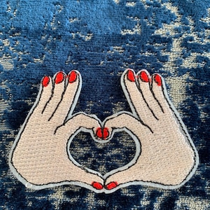 May include: A white embroidered patch of two hands forming a heart shape with red nail polish.