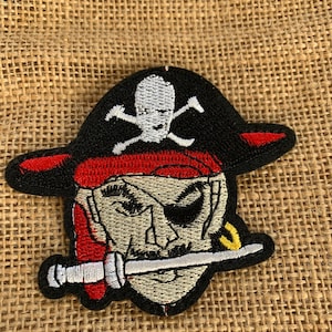 May include: Embroidered pirate patch featuring a skull and crossbones on a black hat with a red trim. The pirate has an eye patch and a sword in its mouth. The patch is on a burlap background.