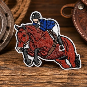 May include: An embroidered patch featuring a chestnut horse jumping over an obstacle with a rider wearing a blue jacket, white breeches, and a black helmet. The horse has black legs and a black tail, and the rider is positioned in a forward seat.