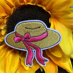 May include: Embroidered patch of a yellow sun hat with a pink ribbon, outlined in white and black. The patch is set against a bright yellow sunflower.