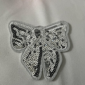 May include: A silver sequined bow applique with a gray felt backing. The bow is filled with shiny silver sequins and outlined with a gray border. The bow is a decorative embellishment for clothing or accessories.