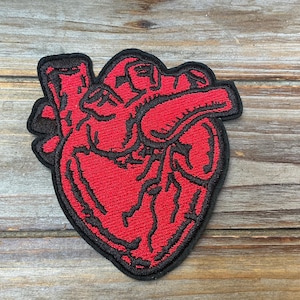 May include: An embroidered patch in the shape of an anatomical human heart. The design features vibrant red thread for the heart muscle and prominent black thread outlining the various chambers, veins, and arteries, finished with a black border.