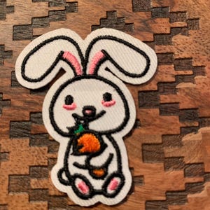 May include: White embroidered bunny rabbit patch with pink ears and a green carrot in its paws. The patch is on a brown wooden background.