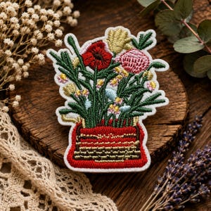 May include: An embroidered patch featuring a red typewriter base with a vibrant bouquet of flowers emerging from the top. The floral arrangement includes a large red poppy, a pink protea, gold accents, and green foliage, all stitched with detailed thread.