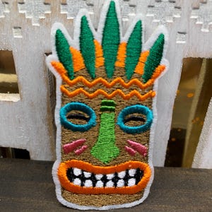 May include: Embroidered patch featuring a Tiki mask design. The mask has a brown background with green, orange, blue, pink, and white accents. The patch has a white border and is likely for clothing or accessories.