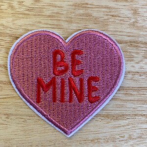 May include: A pink heart-shaped patch with the words "BE MINE" embroidered in red. The patch has a white border and is set against a light wood grain background. This patch is perfect for Valentine's Day or other romantic occasions.