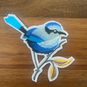 May include: Blue and white embroidered bird patch with yellow leaves on a white background. The bird is perched on a branch.