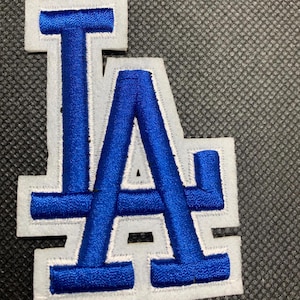 May include: A white felt patch with a blue embroidered "LA" logo. The letters are outlined in white.