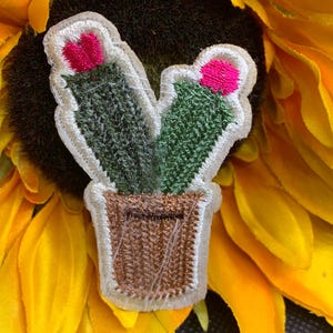 May include: Embroidered cactus patch featuring two green cacti with pink flowers in a brown pot, outlined in white. The patch is set against a yellow sunflower background. This decorative patch is ideal for adding a touch of nature to clothing or accessories.
