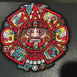 May include: A colorful embroidered patch featuring a detailed Aztec calendar design. The patch has a black border and showcases a central face surrounded by intricate patterns in red, yellow, blue, and white. The design is circular with a hexagonal outer shape.