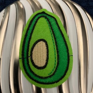 May include: Green felt avocado patch with a brown felt seed inside. The patch has a black outline and green stitching around the edges.