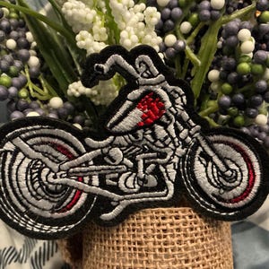 Puede incluir: Parche bordado con un dise&ntilde;o de motocicleta. El parche tiene un borde negro con un dise&ntilde;o de motocicleta plateado y rojo. La motocicleta tiene una apariencia de esqueleto. El parche se coloca en un recipiente envuelto en arpillera con flores artificiales.