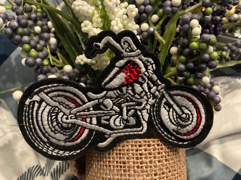 Puede incluir: Parche bordado con un dise&ntilde;o de motocicleta. El parche tiene un borde negro con un dise&ntilde;o de motocicleta plateado y rojo. La motocicleta tiene una apariencia de esqueleto. El parche se coloca en un recipiente envuelto en arpillera con flores artificiales.