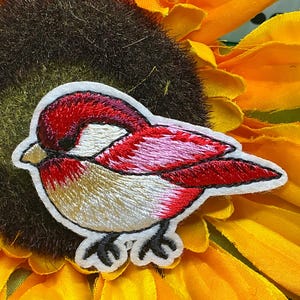 May include: An embroidered patch featuring a small bird with a red head, back, and wings, and a cream-colored chest. The bird has a small gold beak and black feet, outlined in white thread, showcasing detailed stitching throughout the design.