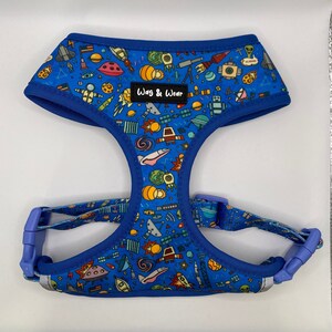 May include: Blue dog harness with a space-themed print featuring rockets, planets, stars, and aliens. The harness has a blue adjustable strap and a black label that reads "Wag & Wear".