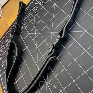 May include: A black leather strap featuring two adjustable loops at each end, secured with black metal snap buttons and a central black plastic buckle mechanism. The strap is positioned over a cutting mat with visible inch markings.