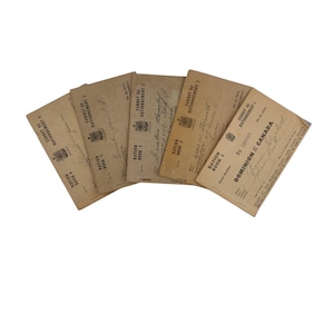 May include: A collection of vintage ration books, fanned out against a white background. The books are tan with black text, including "Ration Book" and "Carnet de Rationnement." Some books have handwritten signatures and stamps.