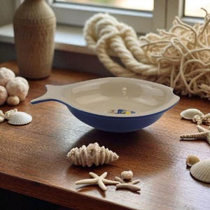 May include: A fish-shaped ceramic dish with a white interior and a blue rim. The dish is on a wooden surface, surrounded by seashells, starfish, and coral. A decorative vase and rope are in the background.