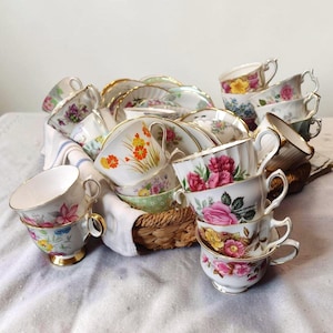 Puede incluir: Una colección de tazas de té y platillos vintage con diseños florales, apilados y dispuestos en una cesta de mimbre. Las tazas presentan bordes y asas doradas, con varios patrones florales en rosa, amarillo y naranja.