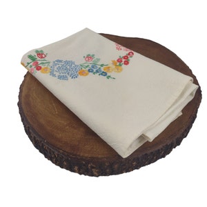 Op de afbeelding: Een witte linnen servet met geborduurde bloemenmotieven in tinten blauw, geel en roze, gevouwen en geplaatst op een ronde bruine houten schijf.
