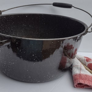 Pode incluir: Uma panela grande de esmalte preto com um acabamento branco salpicado. A panela tem uma alça de arame com uma pega de madeira e uma alça lateral. A panela está sentada sobre uma superfície branca ao lado de uma toalha de cozinha xadrez vermelha e branca e uma colher de pau.