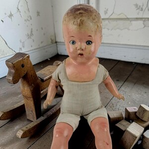 Puede incluir: Una muñeca antigua con rostro pintado, ojos azules y un mono de color neutro. Un caballito balancín de madera y bloques de madera en el fondo sugieren un tema de juguetes vintage.