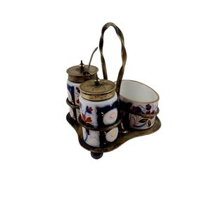 May include: A vintage condiment set with two lidded jars and a small bowl, decorated with white, blue, red, and gold floral patterns. The set is held in a brass-toned metal carrier with a handle.